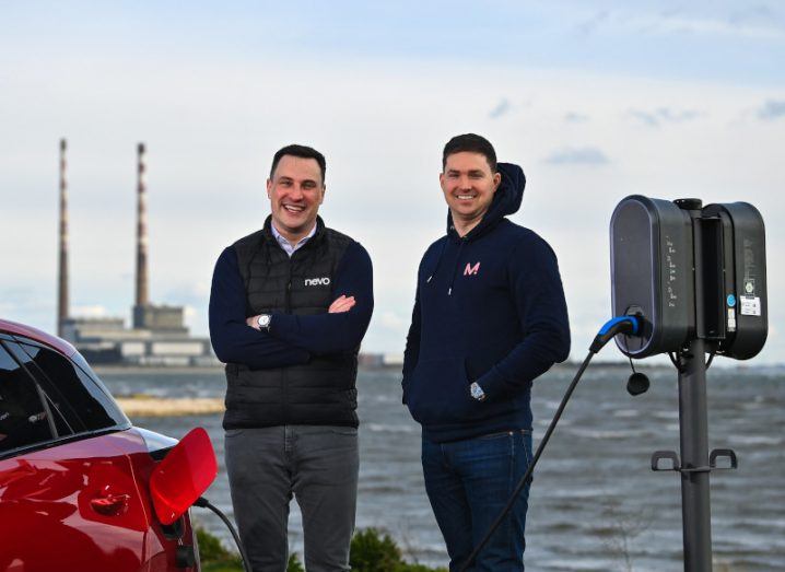 Two men standing next to a red electric car, being charged by a grey charging point. The sea is in the background.