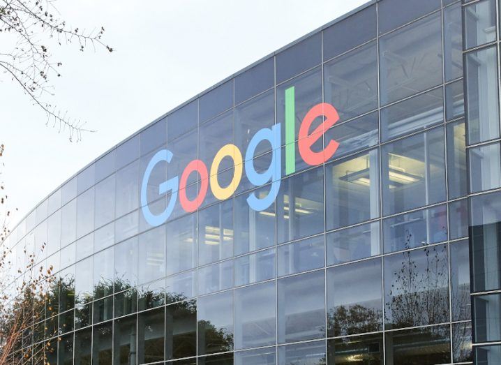 The Google logo on the side of a windowed building, with trees reflecting off the windows and a grey sky above. The company is owned by Alphabet.