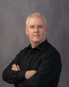 A man with very short white hair looks at the camera standing slightly turned with his arms folded. He is wearing a black shirt.