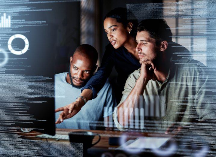 A woman stands behind two men showing them something on a computer by pointing. There are code overlayed on the screen. It is a dark room.