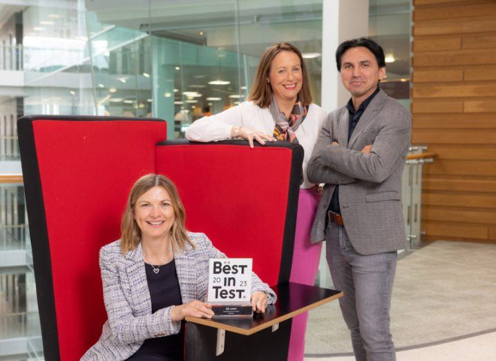 A woman sitting down with a piece of paper that says 'best in test' on it. A man and a woman are standing behind the table, with an office interior in the background. They are part of Vodafone Ireland and Accenture.