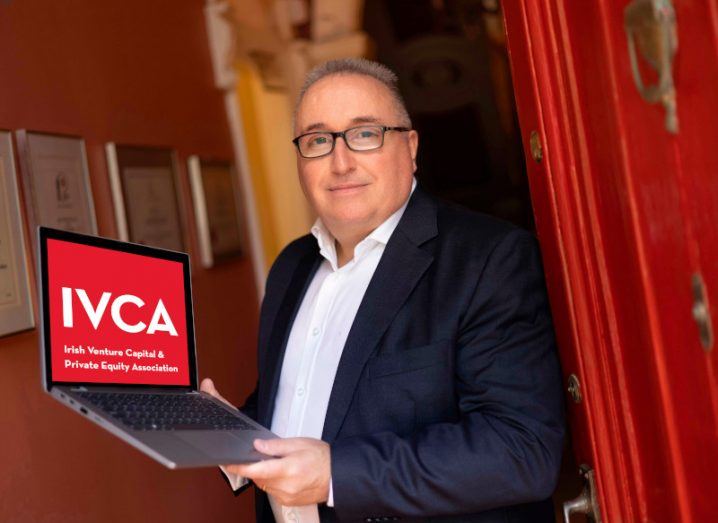 A man wearing a suit and glasses stands holding a laptop displaying the IVCA logo. He is Leo Hamill, chairperson of IVCA.