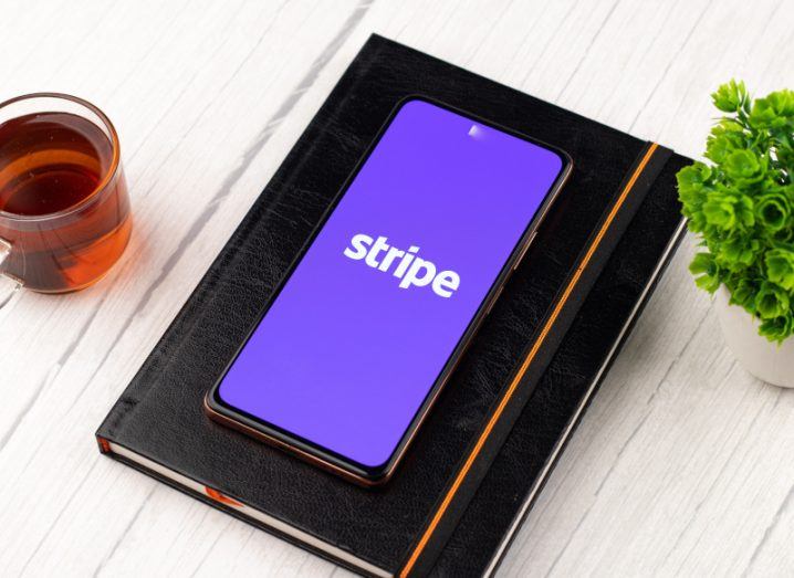 The Stripe logo on a smartphone screen, which is laying on a black book on a wooden surface next to a small plant and a glass with liquid inside it.