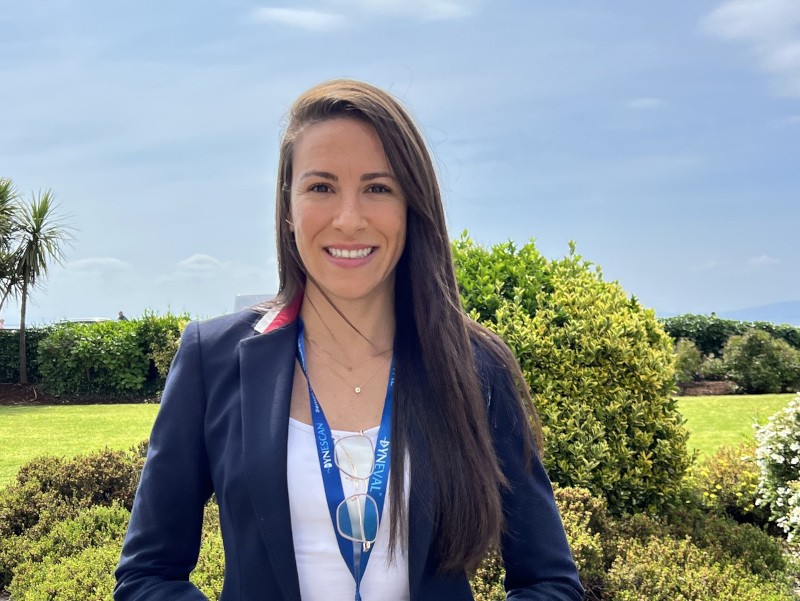 A woman with long, dark hair wears a dark blazer and a white top and smiles at the camera. There are shrubs, a tended lawn and blue skies behind her.