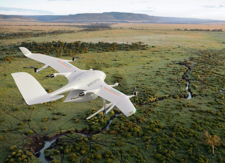 A Wingcopter drone flying over a green landscape with a river visible and mountains in the distance. The Siemens Healthineers logo is visible on the drone.