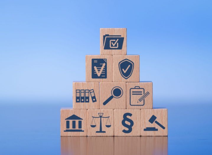 A stack of wooden blocks with various symbols that represent compliance, including folders with check boxes and scales of justice.