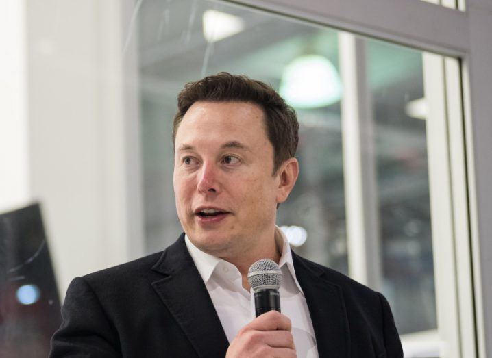 Elon Musk speaking with a microphone in his hand, in a building with a window behind him.