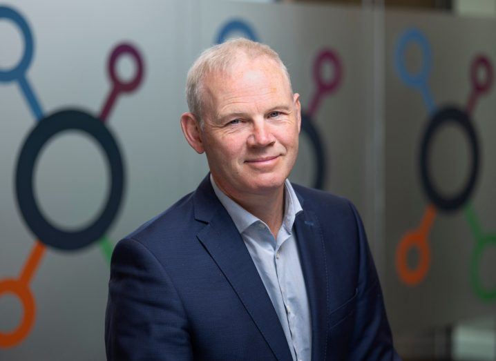 Headshot of a man wearing a navy coloured suit in an indoor space. SuperNode logo on the wall behind him.