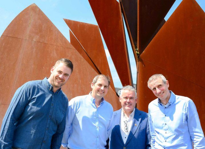 Four men stand next to each other and smile in front of a fountain in Eyre Square, Galway.