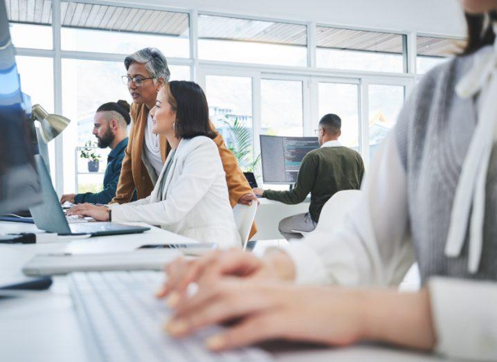 A group of men and women working in an office, with one person typing on a keyboard and someone looking at a computer screen in the background.