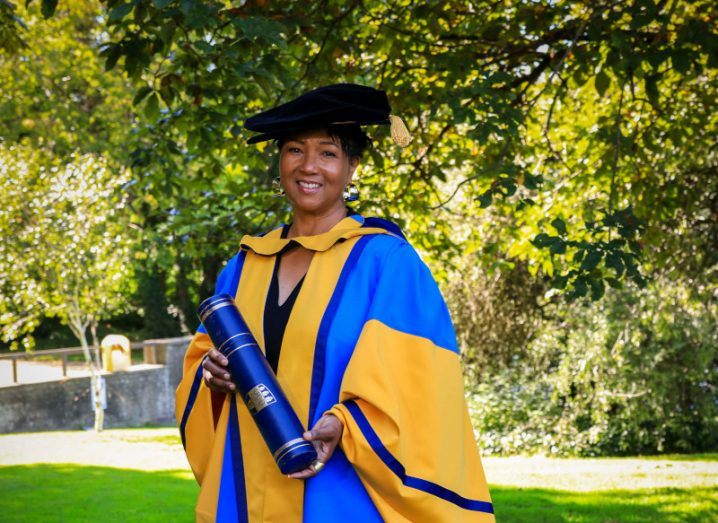 A woman in a blue and yellow graduation ceremony outfit, holding a degree in her hands. She is Dr Mae Carol Jemison