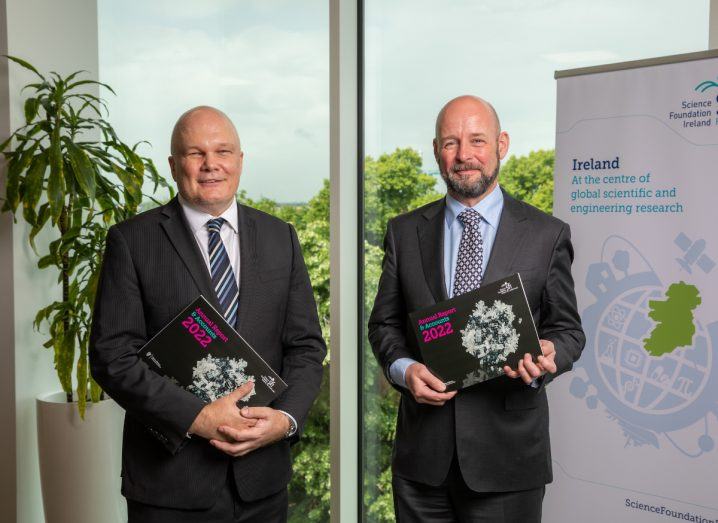 Two men holding the SFI annual report in their hands in front of a poster that has the SFI logo on it.