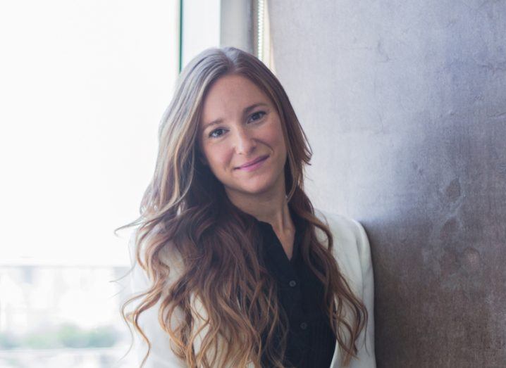 A woman with long brown hair smiles at the camera while leaning against a grey wall. She is wearing a white blazer. She is Professor Cecilia Danesi, an expert in AI ethics and law.