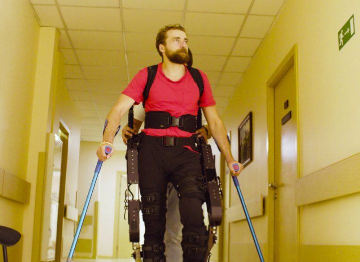 A man holding a pair of crutches while walking with an exoskeleton on his body and a person standing behind him.