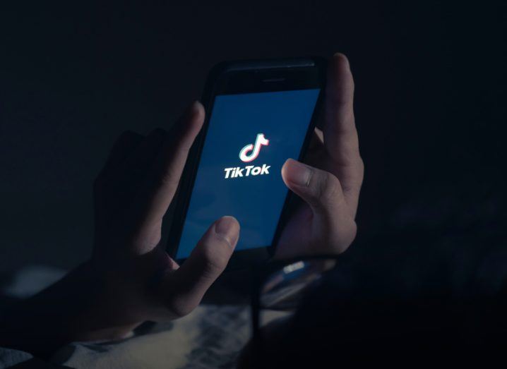 The TikTok logo on a smartphone screen. The phone is in a person's hands in a dark background.