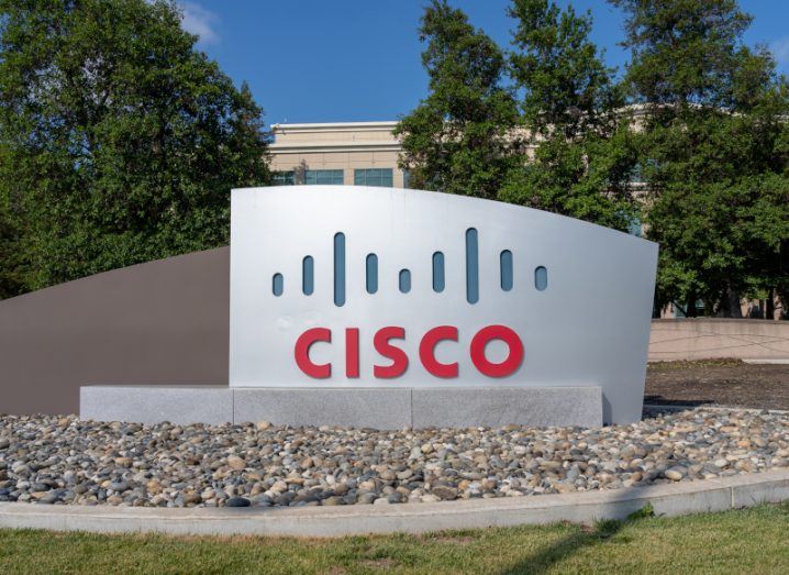 The Cisco logo on a grey sign on a small courtyard, with trees and a building in the background.