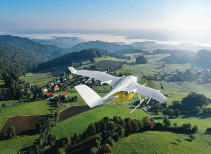 A white drone with the Wingcopter logo on the side, flying above green fields with trees and buildings visible below. A river and a blue sky are visible in the distance.