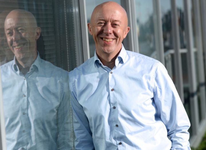 A man in a light blue shirt leans against a window smiling at the camera. He is Version 1 CEO Tom O'Connor