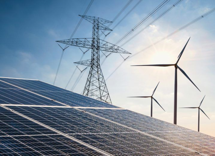 An image to represent the energy sector with solar panels in the foreground and wind turbines and an electricity pile on in the background against a blue sky.