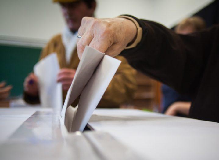 A person putting a piece of paper into a ballot box, with another person holding a piece of paper in the background. Used for the concept of elections and political advertising.