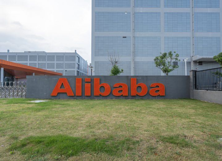 The Alibaba logo on a grey sign on a small patch of grass, with buildings in the background.