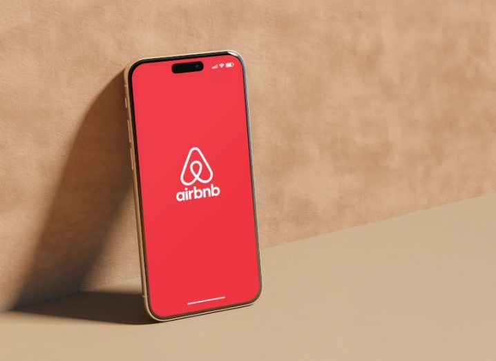 The Airbnb logo on the screen of a smartphone, which is laying next to a brown wall and on a brown floor.