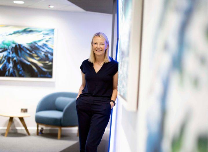 A woman in a black outfit standing by a wall in a room, with a blue chair, a wooden table and a painting in the background. She is Catherine Doyle of Dell Technologies Ireland.