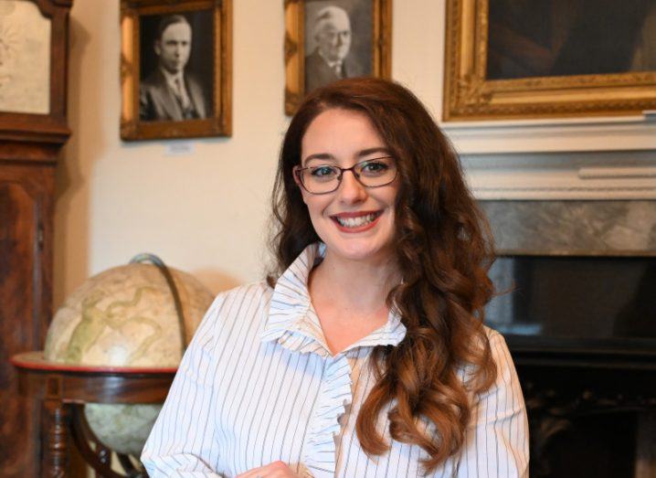 Dr Erin Higgins stands and smiles at the camera in a room with an ornate fireplace and old portraits and an old-fashioned globe behind her. She has long, curly brown hair and glasses and wears a white shirt.