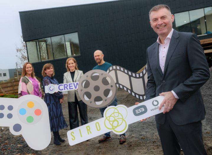 Three women and two men standing in front of a black building, holding multiple paper signs including a game controller, a film reel, a Crew logo and a Siro, ESB and Vodafone sign.