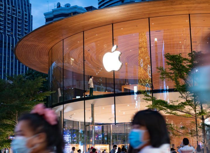 Apple logo on a glass building with people wearing masks walking in the foreground.