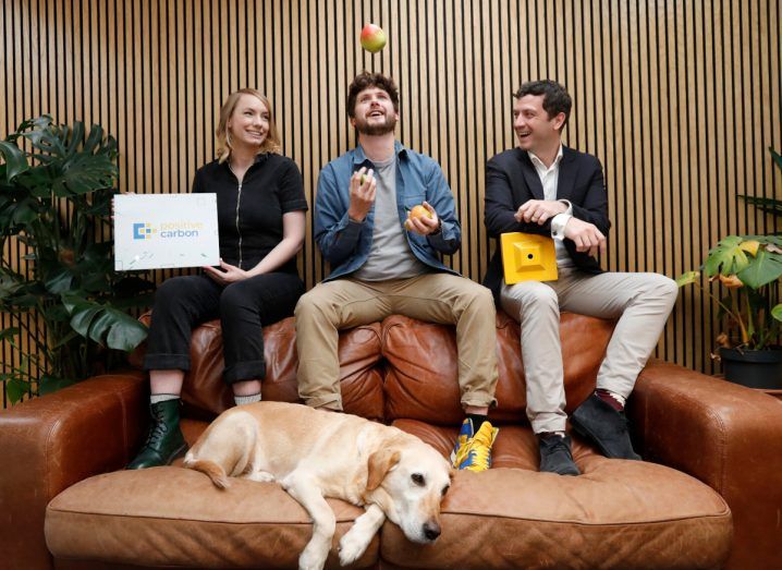 Two men and a woman sit on a sofa while a dog lies in front of them.