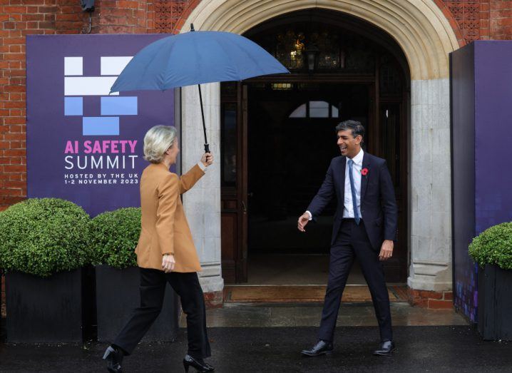 A woman holding an umbrella walks towards a man sticking his hand out in front of an entrance.