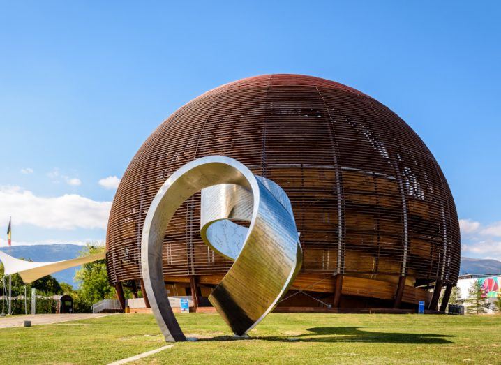 A grey statue in the shape of a ribbon, with a brown circular building behind it. The building is the globe of science and innovation at CERN.