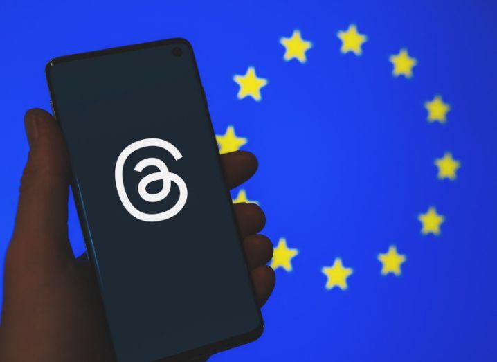 The Threads app logo on a smartphone screen. The phone is held in a person's hand and the EU flag is in the background.
