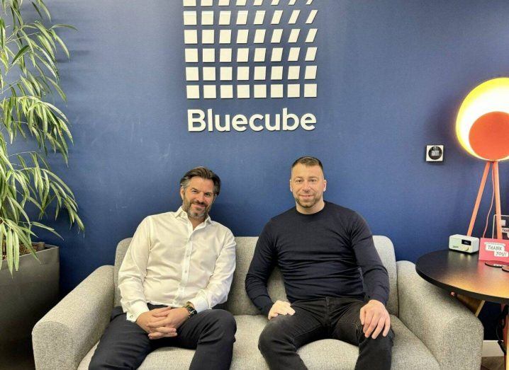 Two men sit on a sofa and pose for a photograph. The Bluecube logo is on a wall behind them.