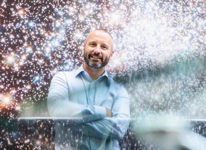 A man in a blue shirt with his arms crossed, standing in front of a wall that has multiple lights of different colours that resembles space. He is Brad Wrigley, the CEO of Varadis.