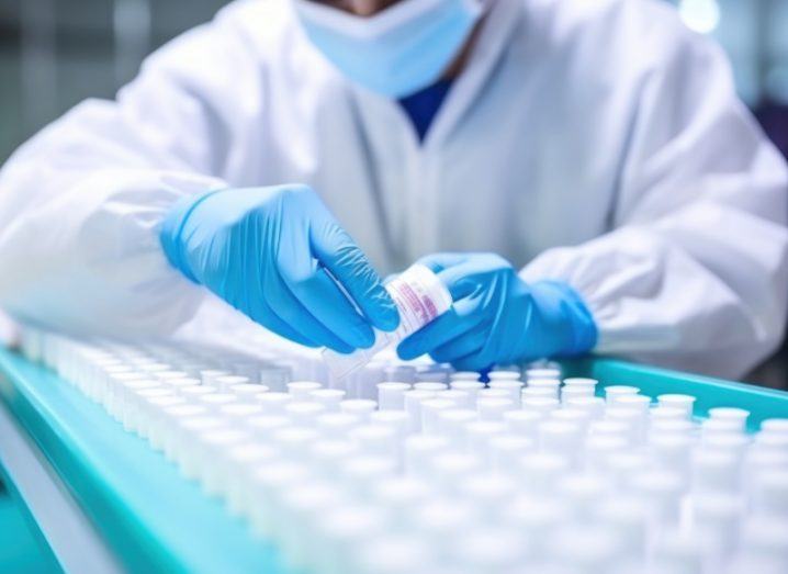 A person wearing a lab coat, blue gloves and a face mask, holding a small container above a large group of containers.