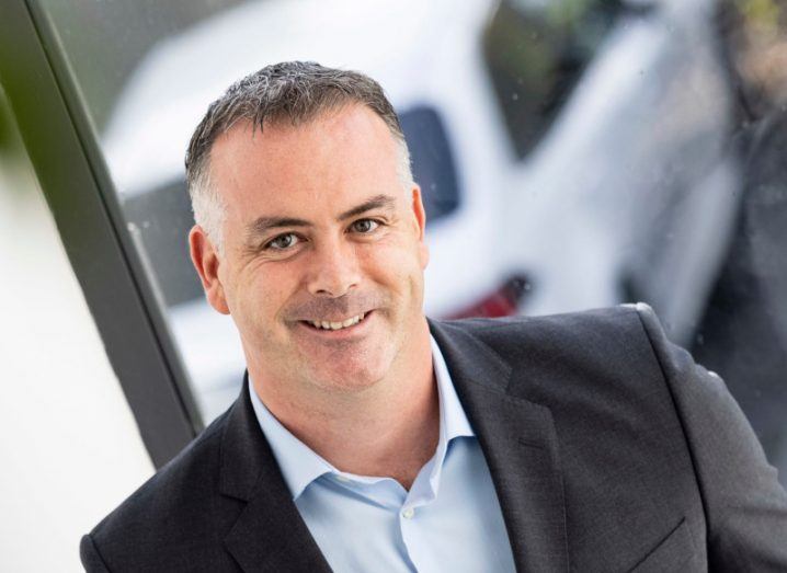 John Delves wearing a black suit and standing in front of a window with a white car in the background.