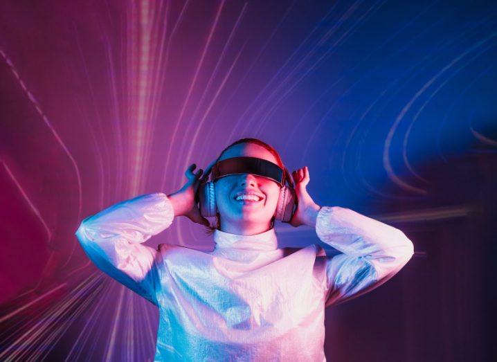 Portrait of a smiling dancing woman in futuristic sunglasses and headphones in pink and blue neon light.