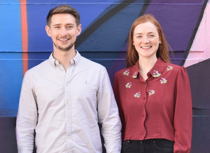 Michael Guerin and Adele Keane stand next to each other in front of a colourful wall with geometric shapes on it. Adele wears a red blouse with small flowers on it and has long red hear. Michael wears a white shirt and had short brown hair and stubble.