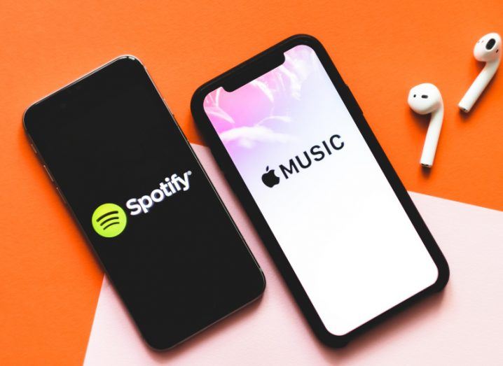 Two smartphones with the Spotify and Apple Music logos on their screens. The phones are laying on a white and orange surface, next to a pair of earphones.