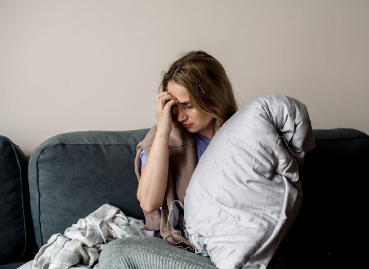 A woman sitting on a chair holding her forehead in pain with one hand, with a pillow and blanket next to her. Used to show the concept of long Covid symptoms.