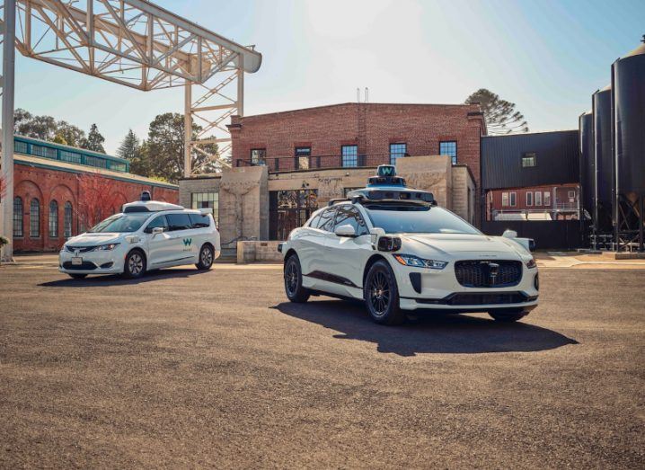 Two Waymo vehicles in front of multiple grey and brown buildings.