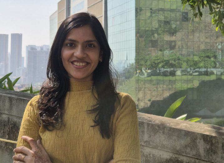 Dhrati Patangia stands with her arms folded and there is some henna visible on her right hand. She is outside in front of greenery and a large glass building on a sunny day.