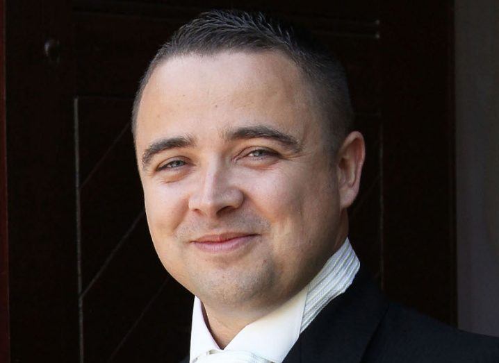 A headshot of Noel Carroll wearing an expensive-looking black suit with white shirt and tie, against a dark background.