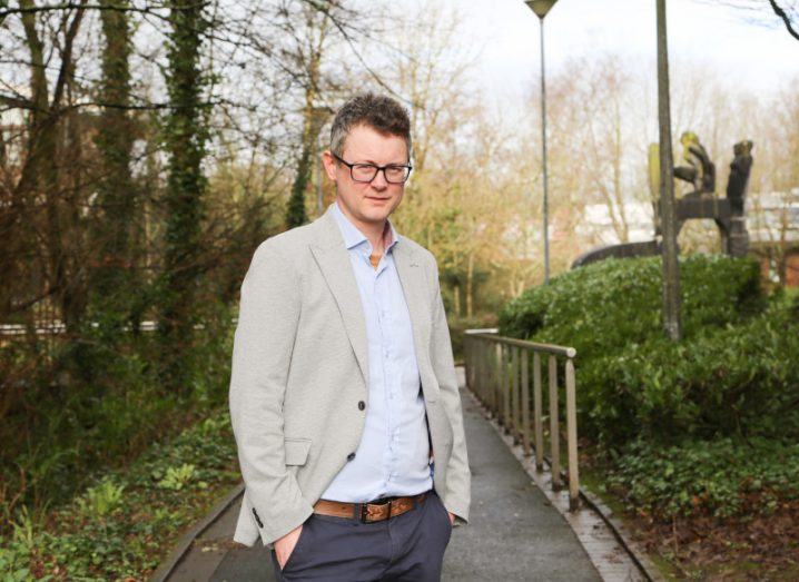 A photo of Prof Maurice Collins from UL standing outside in a wooded area on a concrete path. He wears a light blazer, blue shirt and dark trousers and has his hands in his pockets.