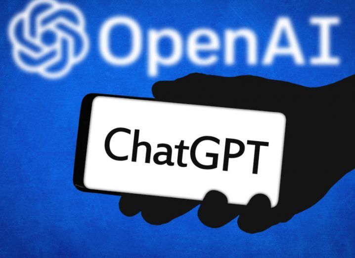 The ChatGPT logo on the screen of a smartphone, held in a person's hand with the OpenAI logo behind it on a blue background.