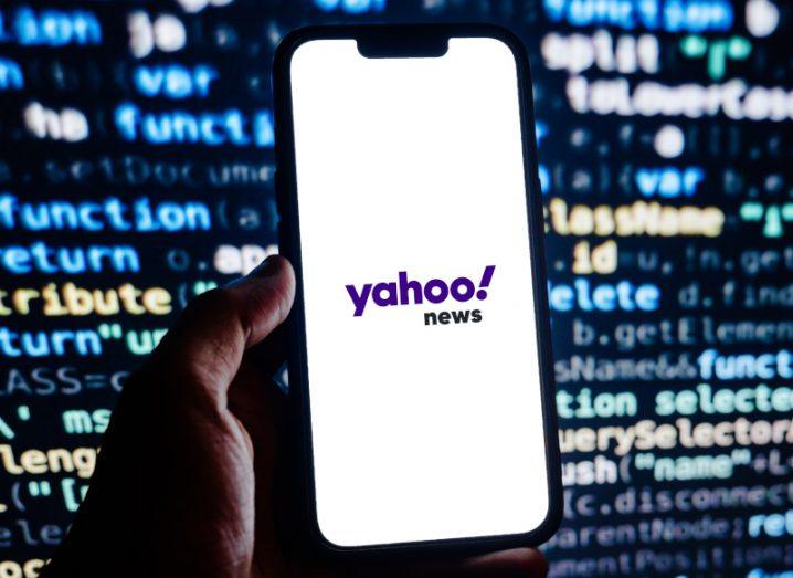 A hand holding a phone with the Yahoo News logo on its screen with a screen of code in the background.