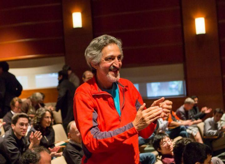 A man in a red jacket and jeans stands in in a crowd of seated people in a lecture hall applauding. He is Avi Wigderson.