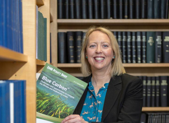 Dr Fiona Grant standing in front of rows of books and holding a book by the European Marine Board.